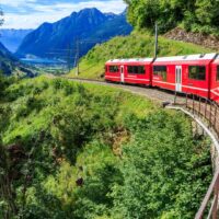 A red Swiss train curves along a mountain track surrounded by green hills.