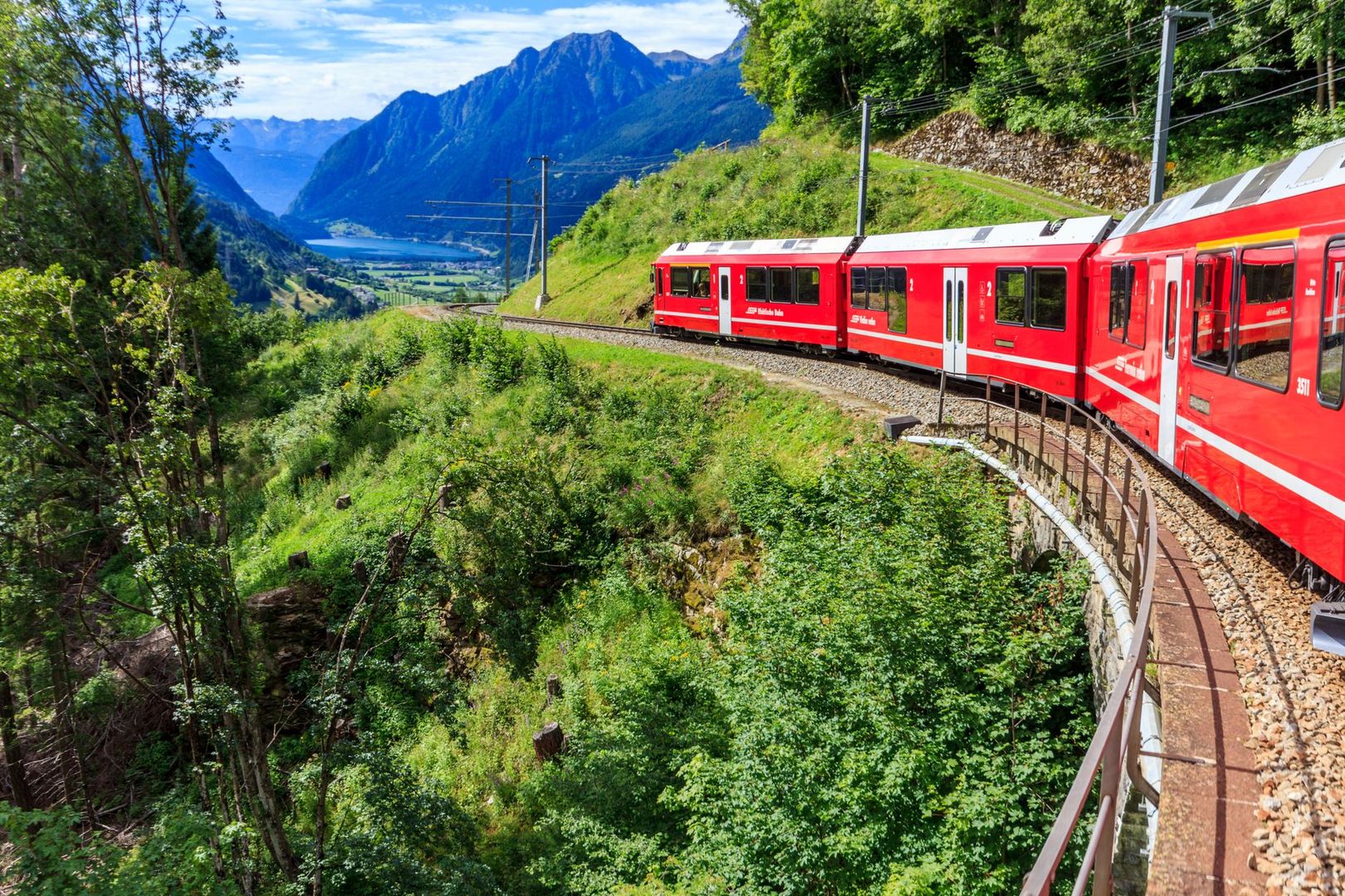 A red Swiss train curves along a mountain track surrounded by green hills.